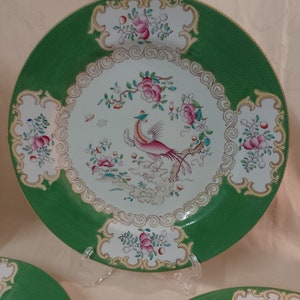 May include: A set of three green and white ceramic dinner plates with a gold trim. The center of each plate features a pink and white bird with a long tail, surrounded by floral designs.