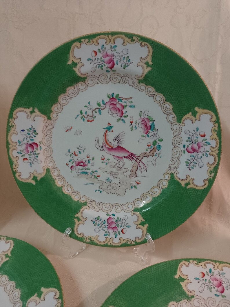 May include: A set of three green and white ceramic dinner plates with a gold trim. The center of each plate features a pink and white bird with a long tail, surrounded by floral designs.