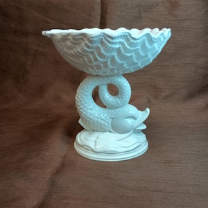 Puede incluir: Tazón de cerámica blanca con borde festoneado y superficie texturizada. El tazón está sostenido por una figura de delfín de cerámica blanca.