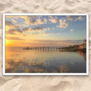 May include: A framed photograph of a sunset over a calm body of water with a wooden pier extending into the water. The sky is a vibrant orange and pink, with fluffy white clouds. A small building is visible on the shore.