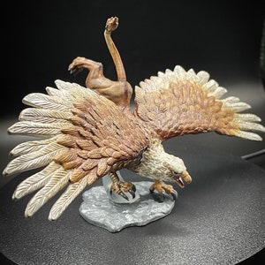May include: A brown and white painted miniature figurine of a griffin, a mythical creature with the head and wings of an eagle and the body of a lion. The griffin is posed in mid-flight with its wings spread wide and its talons extended.