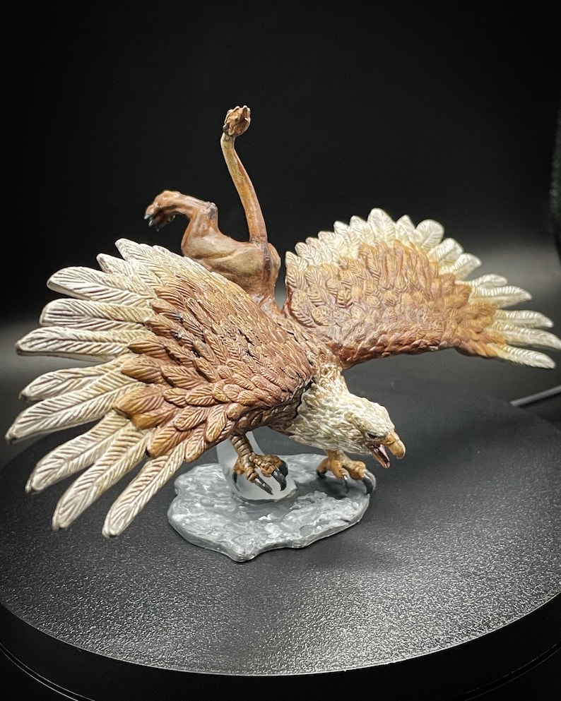 May include: A brown and white painted miniature figurine of a griffin, a mythical creature with the head and wings of an eagle and the body of a lion. The griffin is posed in mid-flight with its wings spread wide and its talons extended.