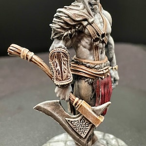 May include: A painted miniature figurine of a warrior holding a double-bladed axe. The figure is wearing a brown leather armor with fur trim and a red loincloth. The axe has a silver blade and a brown wooden handle.