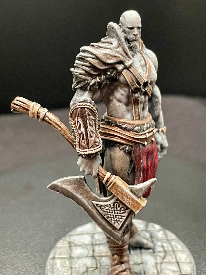 May include: A painted miniature figurine of a warrior holding a double-bladed axe. The figure is wearing a brown leather armor with fur trim and a red loincloth. The axe has a silver blade and a brown wooden handle.