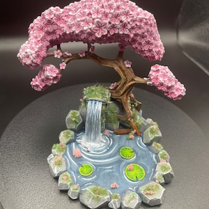 May include: A miniature ceramic scene of a pink cherry blossom tree with a waterfall flowing into a pond with lily pads. The base is made of gray rocks and green moss.