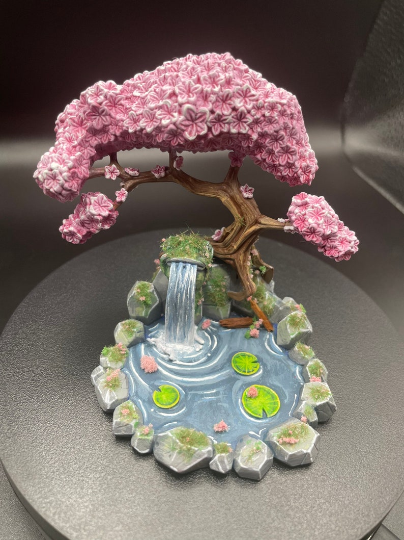 May include: A miniature ceramic scene of a pink cherry blossom tree with a waterfall flowing into a pond with lily pads. The base is made of gray rocks and green moss.