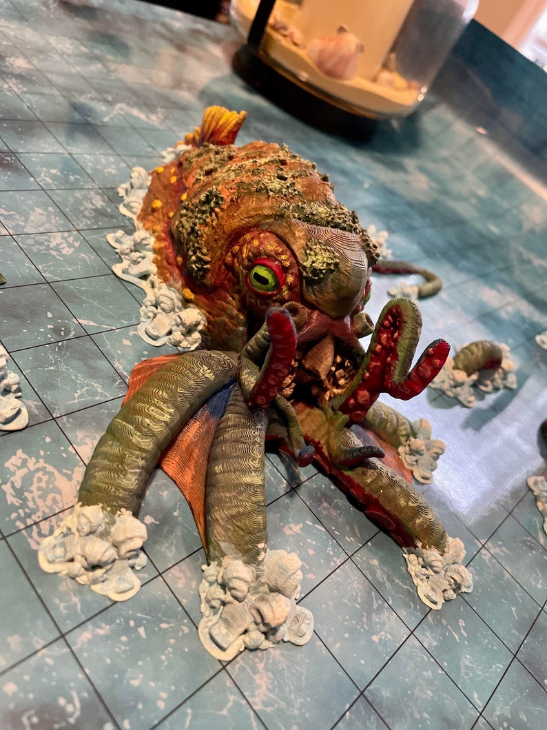 May include: A painted miniature of a green, red, and orange octopus with a large, open mouth and one visible green eye. The octopus is surrounded by white foam-like material, suggesting it is in water.