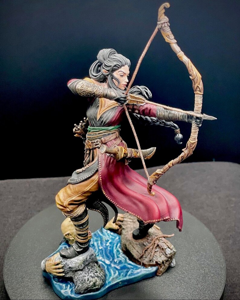May include: A painted miniature figurine of a female elf archer, standing on a rocky base with water flowing around her feet. She is wearing a red and gold outfit and holding a bow and arrow.