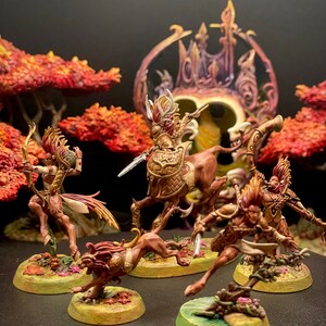 May include: A set of five miniature figures painted in shades of brown, gold, and white. The figures are posed in a dynamic action stance, with some holding weapons. The figures are set against a backdrop of a dark background and a red and orange tree.