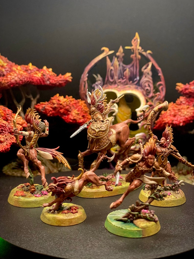 May include: A set of five miniature figures painted in shades of brown, gold, and white. The figures are posed in a dynamic action stance, with some holding weapons. The figures are set against a backdrop of a dark background and a red and orange tree.
