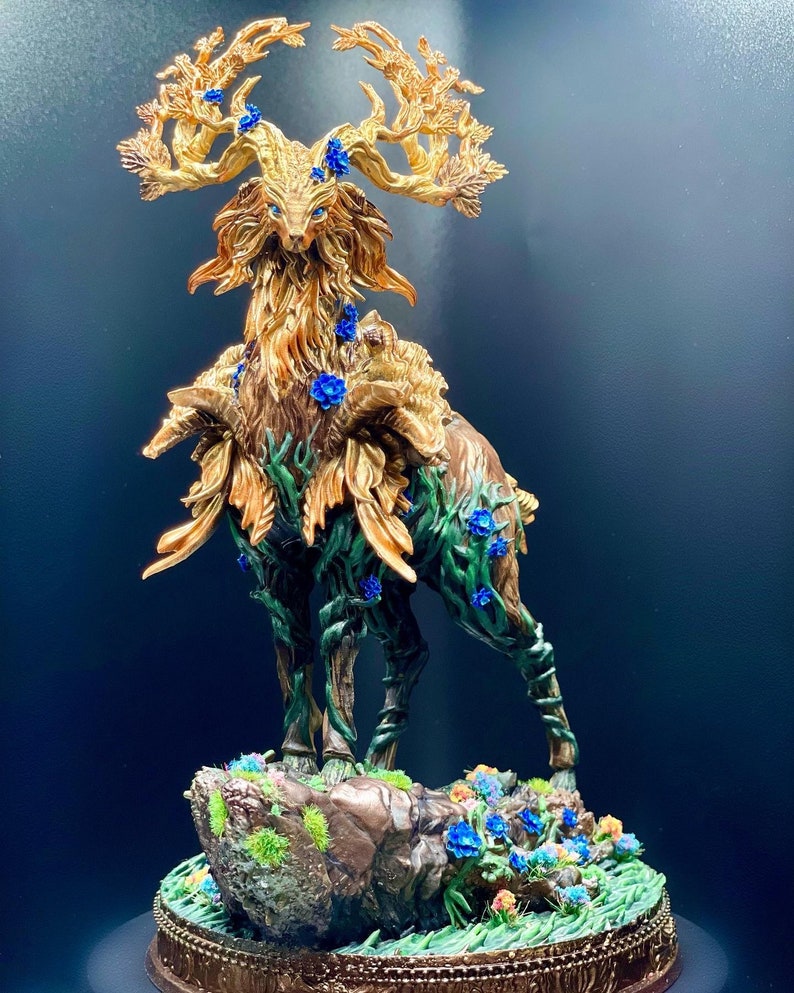 May include: A golden deer statue with antlers made of branches and leaves. The deer is covered in vines and blue flowers. The statue is standing on a rock base with green moss and flowers.