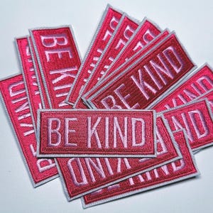 Może przedstawiać: Stos różowych łat z białą nicią. Każda łata ma wyszyte słowa "Be Kind".