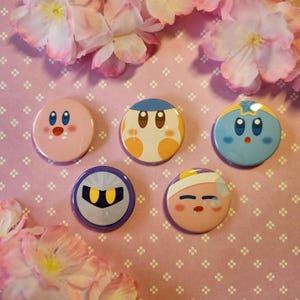 Peut inclure: Collection de cinq badges ronds représentant des personnages. Les badges présentent divers personnages de dessins animés dans des tons de rose, de bleu, d'orange et de violet. Le fond est une surface rose avec des motifs floraux blancs et des fleurs roses.