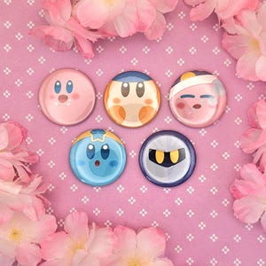 Peut inclure: Cinq badges ronds représentant des visages de personnages de la série de jeux vidéo Kirby, dont Kirby, Waddle Dee et Meta Knight, disposés en groupe sur un fond rose avec un motif de grille blanche.