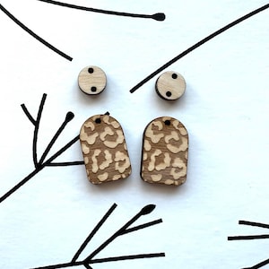 Puede incluir: Un par de pendientes de madera con diseño de estampado de leopardo. Los pendientes son de forma rectangular y tienen un pequeño poste redondo de madera para sujetarse a la oreja.