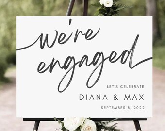 May include: White sign with black calligraphy that reads "we're engaged" with the names "Diana & Max" and the date "September 3, 2022" below. The sign is on an easel with white flowers.