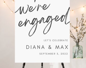 May include: A white sign on a black tripod stand with the words "We're engaged" in a handwritten style. Below, it reads "Let's Celebrate" followed by "Diana & Max" and the date "September 3, 2022".