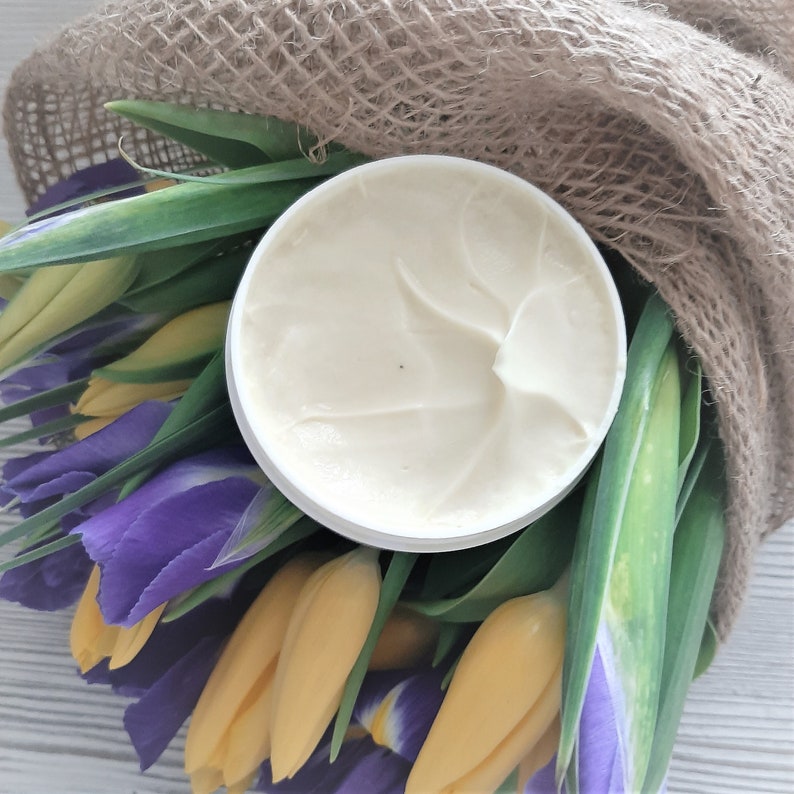 Puede incluir: Un tarro blanco de crema con un dise&ntilde;o de remolino en la parte superior, rodeado de flores amarillas y moradas y una tela de arpillera.