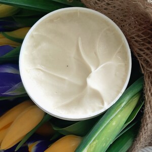 Puede incluir: Un tarro blanco de crema con un dise&ntilde;o de remolino en la parte superior. El tarro est&aacute; rodeado de flores amarillas y moradas.