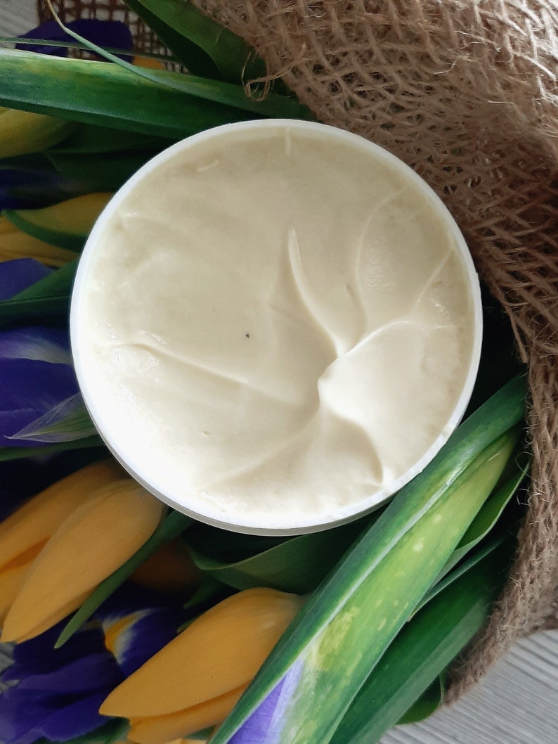 Puede incluir: Un tarro blanco de crema con un dise&ntilde;o de remolino en la parte superior. El tarro est&aacute; rodeado de flores amarillas y moradas.