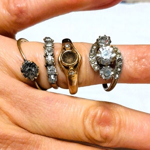Op de afbeelding: Een hand met vier ringen. De ringen zijn allemaal in verschillende stijlen en hebben verschillende edelstenen. De ringen zitten aan de wijsvinger en middelvinger.