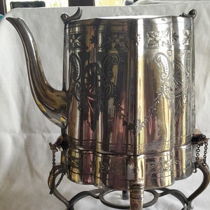 Op de afbeelding: Zilveren theepot met sierlijke bloemen- en bladgravures. De theepot heeft een tuit, een handvat en een decoratieve voet met een warmhoudstandaard. Het ontwerp omvat een ketting en een deksel. De theepot is ongeveer 30 cm hoog.