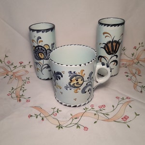 Könnte beinhalten: Drei Keramikbecher mit blauen und gelben Blumendesigns. Die Becher stehen auf einer weißen Tischdecke mit rosa Blumen und einem hellrosa Band.