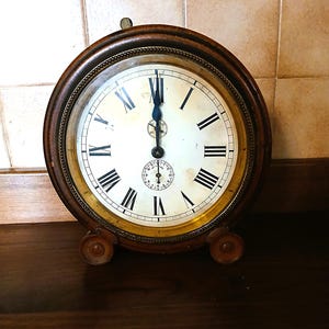 Op de afbeelding: Antieke ronde klok met een donkerhouten frame en een witte wijzerplaat met zwarte Romeinse cijfers. De klok heeft een kleinere subwijzerplaat en decoratieve voetjes. De wijzers staan op twaalf uur. De klok staat op een donkerhouten oppervlak.