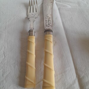 Könnte beinhalten: Ein Besteckset aus Silber mit Elfenbeinstielen. Das Messer hat ein verziertes Design und die Gabel ein schlichtes Design. Beide Teile haben einen gedrehten Griff.