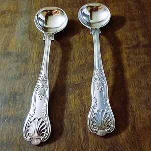 Può includere: Due cucchiai da portata d'argento con manici incisi e ciotole rotonde. I cucchiai hanno una superficie riflettente e sono appoggiati su uno sfondo di legno scuro. I manici presentano un elaborato motivo floreale e a conchiglia.