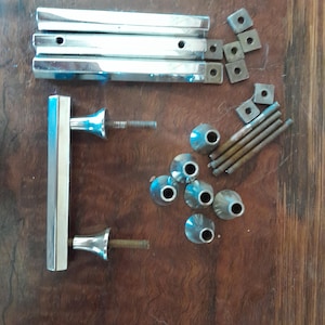 Op de afbeelding: Een assortiment glanzende, zilverkleurige hardwarecomponenten, waaronder deurgrepen, vierkante montageplaten en decoratieve knoppen. Ook aanwezig zijn draadstangen en schroeven, allemaal gerangschikt op een donker, houtnerfoppervlak. Deze items lijken bedoeld te zijn voor meubel- of deurinstallatie.