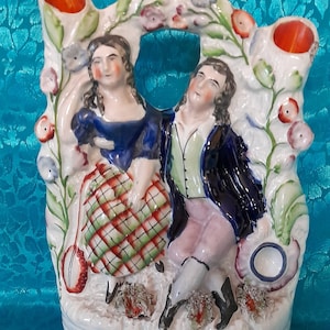 Könnte beinhalten: Eine weiße Keramikfigur eines Paares in einer Gartenlandschaft. Die Frau trägt ein blaues Kleid mit einem rot-grünen karierten Rock. Der Mann trägt eine grüne Weste und eine blaue Jacke. Die Figur hat zwei orangefarbene Blumenhalter an den Seiten.