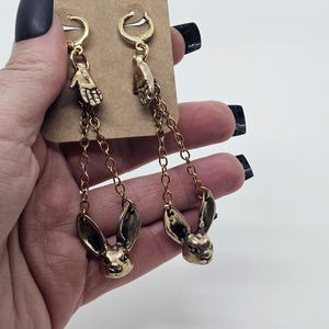 Könnte beinhalten: Goldfarbene Ohrringe mit einem Hand- und Hasenkopf-Design. Die Ohrringe haben einen Klappverschluss und sind an einer filigranen Kette aufgehängt. Die Hasenköpfe sind detailliert mit sichtbaren Ohren und Gesichtszügen.