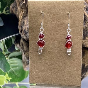 May include: Silver wire earrings featuring three red gemstones. The gemstones are wrapped in silver wire and hang from a silver hook. The earrings are displayed on a brown card.