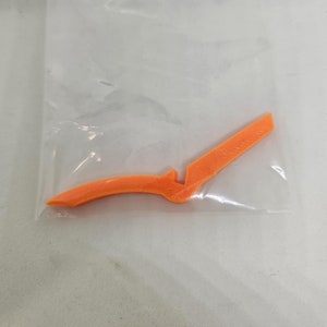 以下が含まれることがあります： 透明なビニール袋に入った、オレンジ色の3Dプリントされた、様式化されたナイフまたは刃のようなオブジェクト。オブジェクトは、湾曲したハンドルと、まっすぐで角度の付いた刃を持っています。ビニール袋は透明です。