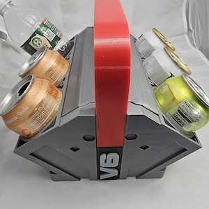 May include: A gray, 3D-printed carrier with a red handle, holding soda cans and a water bottle. The carrier has a V6 marking on the front. The cans are orange and yellow, and the bottle is clear.