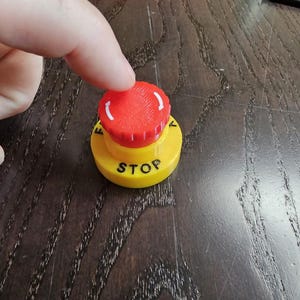 Könnte beinhalten: Ein roter und gelber Not-Aus-Knopf mit dem Wort "STOP" auf der gelben Basis. Der rote Knopf hat weiße Pfeile. Ein Finger drückt den Knopf.