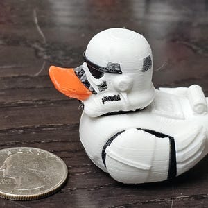 May include: White rubber duck with a Stormtrooper helmet design. The duck has an orange beak and black details on the helmet.