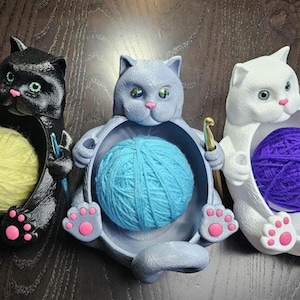 May include: Three cat-shaped yarn holders in black, gray, and white. Each cat holds a ball of yarn in its belly, with a crochet hook in its paw. The black cat has yellow yarn, the gray cat has blue yarn, and the white cat has purple yarn.