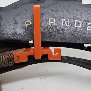 May include: Close-up of a car's gear shift indicator with the letters "P R N D 2" in white. An orange plastic piece is attached to the gear shift mechanism. A metal spring is also visible.
