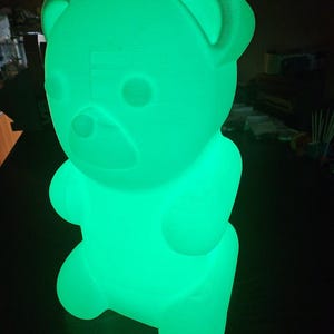 Può includere: Lampada a forma di orsetto verde brillante. La lampada ha un design liscio e arrotondato con orecchie, occhi e naso visibili. La luce emana dall'interno, creando un bagliore soffuso e ambientale. È un oggetto decorativo.