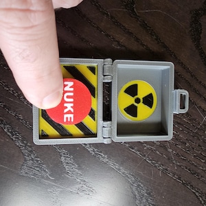 May include: A gray, hinged box with a red button labeled "NUKE" and a yellow and black hazard pattern. The open box reveals a yellow radiation symbol. The item is on a dark wood surface.