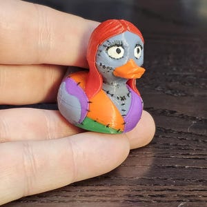 May include: A colorful, 3D-printed rubber ducky featuring a design inspired by Sally from The Nightmare Before Christmas. The duck has a gray face, orange beak, red hair, and a patchwork body in purple, orange, and green.