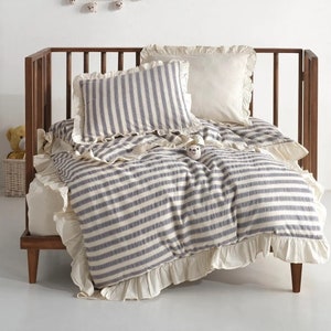 Puede incluir: Una cuna de madera con ropa de cama a rayas azules y blancas, que incluye un edredón y una almohada. La ropa de cama tiene bordes con volantes. Una guirnalda de pequeñas cabezas de oso cuelga sobre la cuna.