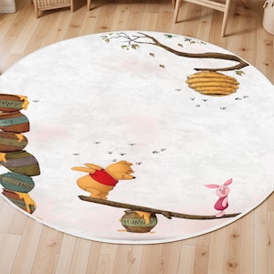 May include: Round area rug featuring a whimsical illustration of Winnie the Pooh and Piglet. The design includes a seesaw with a honey pot, a stack of honey pots, a beehive, and the word "Hunny". The rug has a light pink and white background.