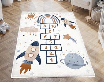 Könnte beinhalten: Ein weißer und blauer Spielteppich für Kinder mit einem Hopscotch-Spiel, einer Rakete, einem Regenbogen, einer Wolke und einem Planeten. Die Zahlen 1 bis 10 befinden sich auf den Hopscotch-Feldern.