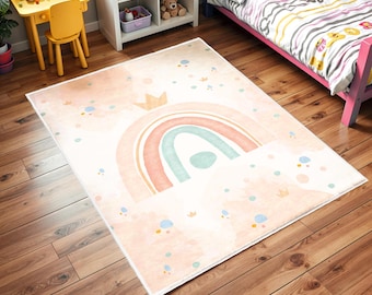May include: A pink and white children's rug with a rainbow design and a crown on top. The rug has a watercolor effect and is decorated with small dots and stars.