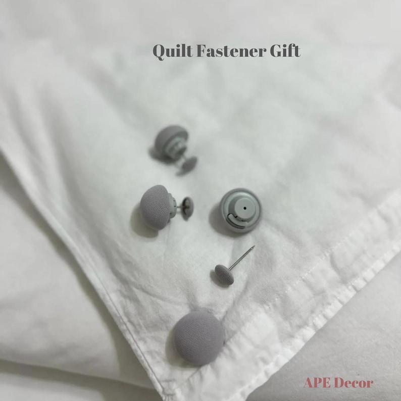 Op de afbeelding: Vijf grijze, met stof beklede knoppen met metalen pinnen. De knoppen liggen op een wit oppervlak. De tekst "Quilt Fastener Gift" staat bovenaan de afbeelding. De tekst "APE Decor" staat onderaan de afbeelding.
