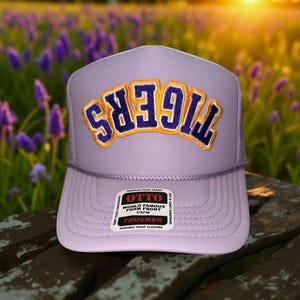 May include: A lavender trucker hat with the word "TIGERS" embroidered in gold and navy blue. The hat has a foam front and a mesh back. The label reads "OTTO WORLD FAMOUS FOAM FRONT TRUCKER DURABLE SNAP CLOSURE".