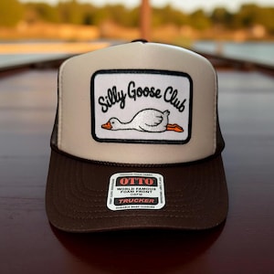 May include: A brown and tan trucker hat with a white front panel featuring a "Silly Goose Club" patch. The patch shows a cartoon goose. The hat has a mesh back and a snap closure. The hat also has a "World Famous Foam Front" tag.